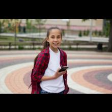 A young girl standing in the park, holding a mobile phone, looking into the camera and smiling